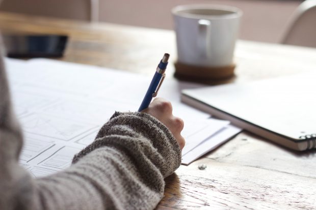 person writing in exercise book, mug and other papers on desk