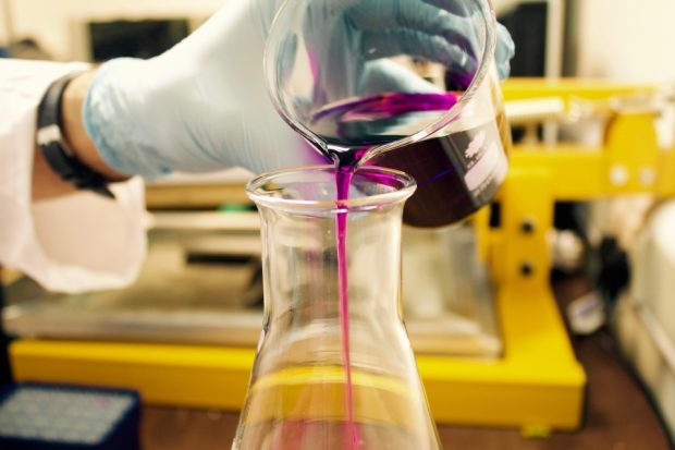 Image of person's hand pouring purple liquid from glass jar into another glass receptacle. Person is wearing rubber safety glove. Only their wrist is visible - they appear to be wearing an overall. Yellow metal equipment of some sort in background (image is shot so that it isn't clear exactly what this is but impression is that it is scientific equipment).
