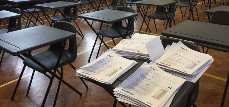 Four piles of exam papers on a desk in an empty exam hall