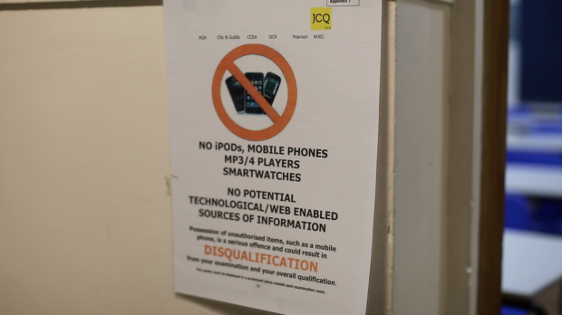 Image shows poster stuck on doorway (inference is this is the entrance to an exam). It shows the JCQ logo in the right hand corner. Below is an image of 3 mobile phones inside a red circle border, with a red line struck through them. Text below reads: No ipods, mobile phones, MP3/4 players, smartwatches. No potential technological/web enabled sources of information. Disqualification. There are 4 additional rows of text but the photo is out of focus and these words cannot be read