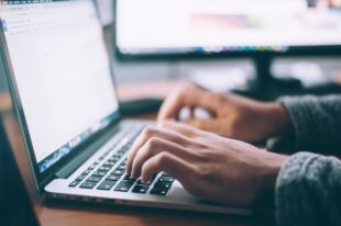 Picture of laptop keyboard and screen with two hands, visible to wrist, typing