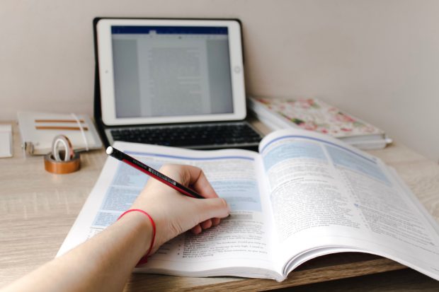 A person making notes with their left hand in a textbook on a desk, in front of a tablet computer.