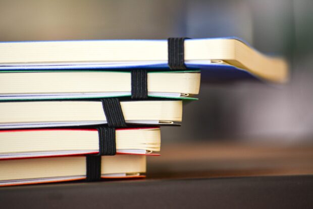 An offset stack of five exercise books, one yellow, one red, one grey, one green and one blue.