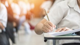 Young person sitting at a small desk using a pencil to write on a notepad, with a window behind them.