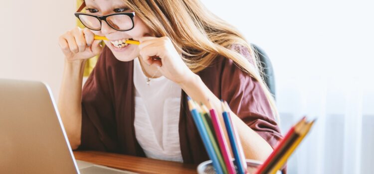 A girl biting into a pencil, looking pleased at something on her computer screen